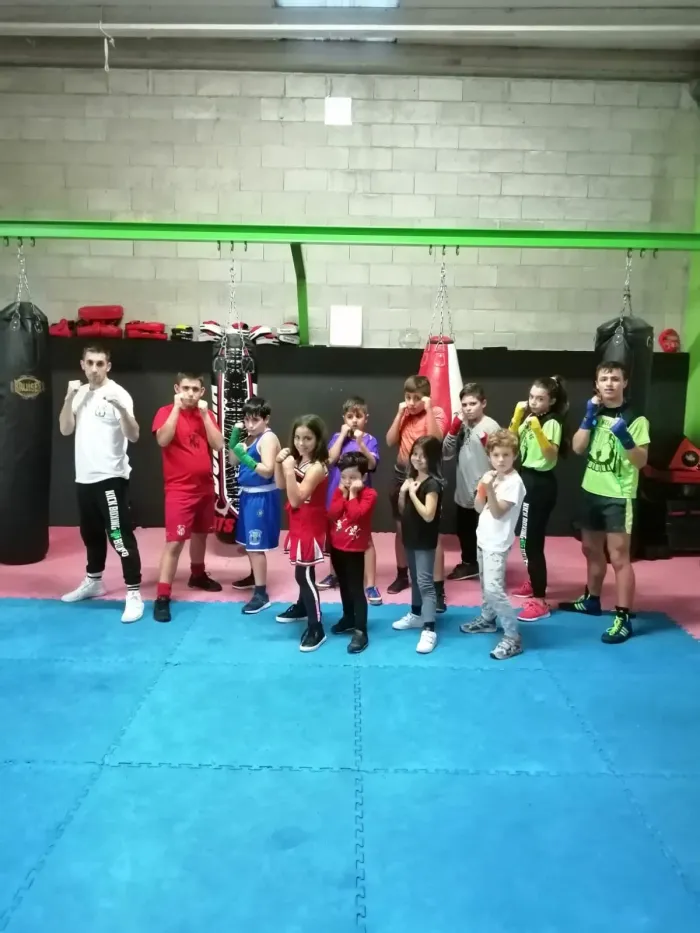 Grupo de niños y niñas del club posando en guardia junto a su entrenador, delante de los sacos del gimnasio
