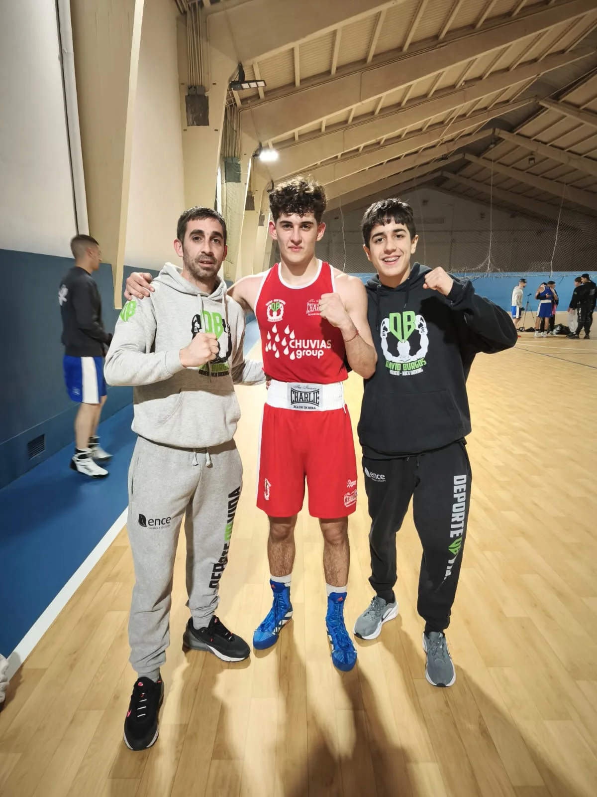 Boxeador del club con equipación roja posando con su entrenador y un compañero en el pabellón de Lugo