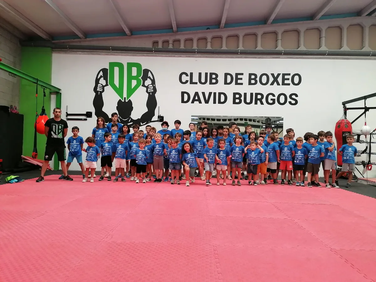 Los niños del campamento Casiña de Teo, con camiseta azul, posando en grupo con el entrenador bajo el rótulo del Club de Boxeo David Burgos